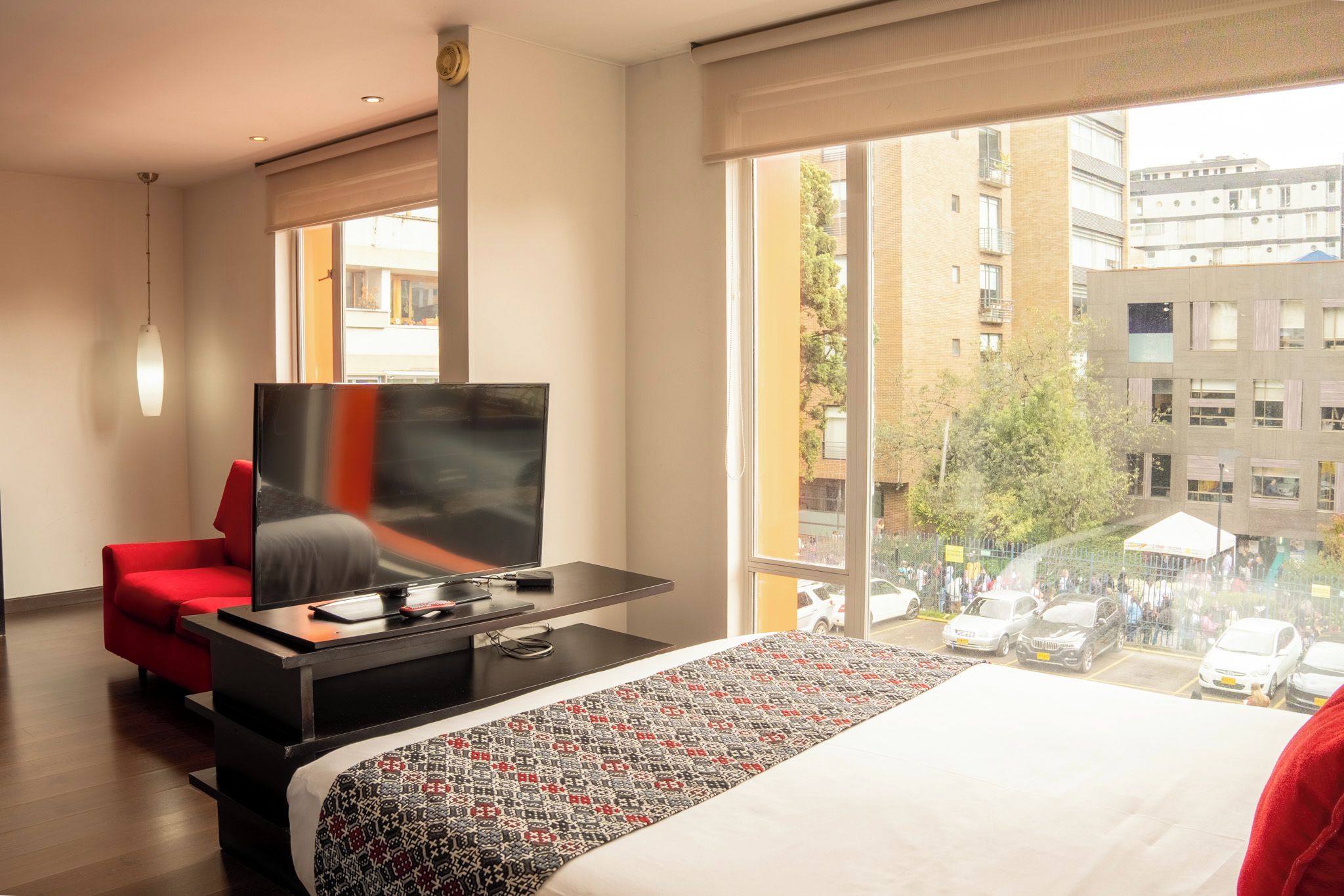 Habitación de hotel con cama, televisión, una silla roja y un ventanal que ofrece vistas de la ciudad.