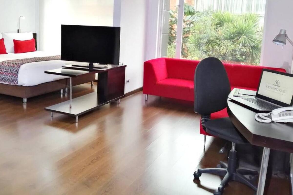 Quarto de hotel moderno com cama, televisão, escrivaninha com um laptop e um sofá vermelho.
