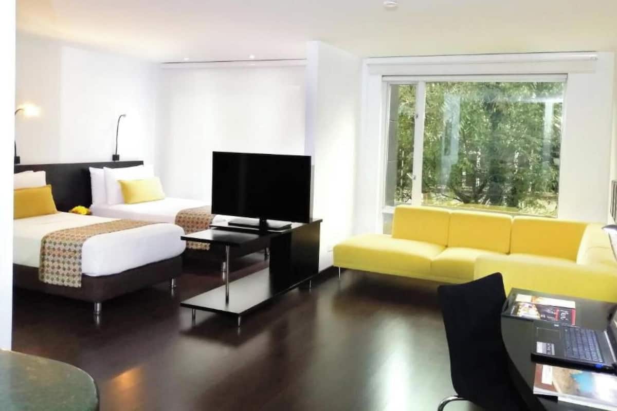 Quarto de hotel moderno com cama, televisão, sofá e secretária com um computador portátil.