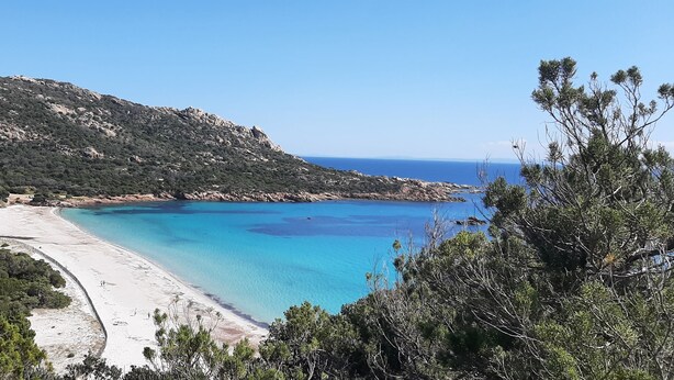 A coastal landscape with a sandy beach, clear blue waters, and a hilly backdrop.