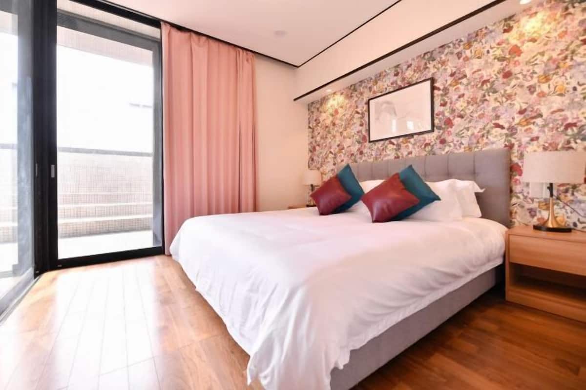 A bedroom with a large bed, a nightstand, a window with curtains, and a floral wallpaper.