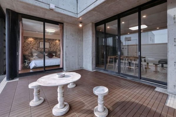 A modern interior with a dining area featuring a round table and stools, a visible bedroom with a bed and curtains, and a large sliding glass door leading to an outdoor seating area. - BRUCE HOUSE (Izumisano)