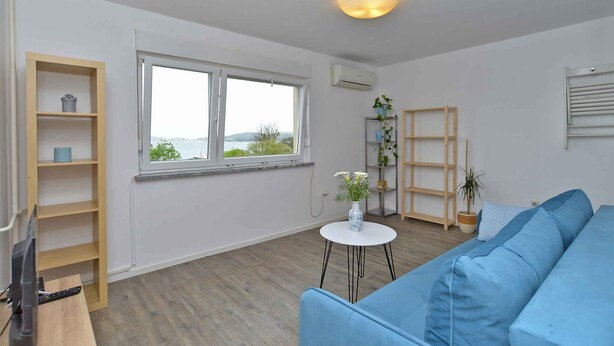 A spacious living room with a large window, a blue sofa, a small round table, and a wooden shelf unit.