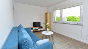 A modern living room with a blue sofa, a small white table with a vase, a TV on a wooden stand, and a large window with a view of greenery.