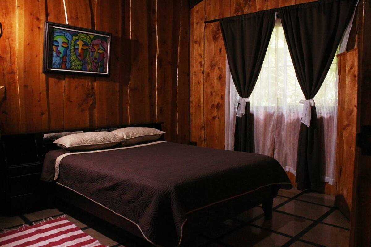A wooden room with a bed, a framed artwork on the wall, and a window with curtains.
