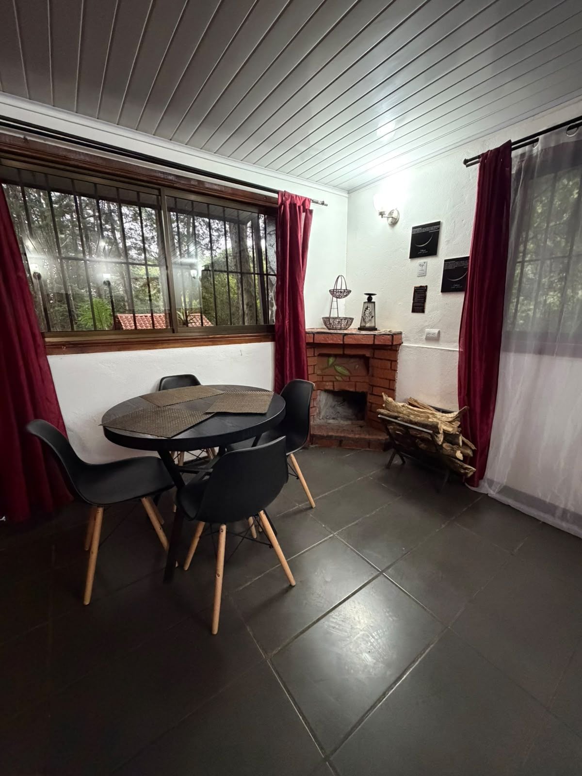 Un comedor con una mesa redonda y cuatro sillas, una chimenea de ladrillo y una ventana con cortinas.