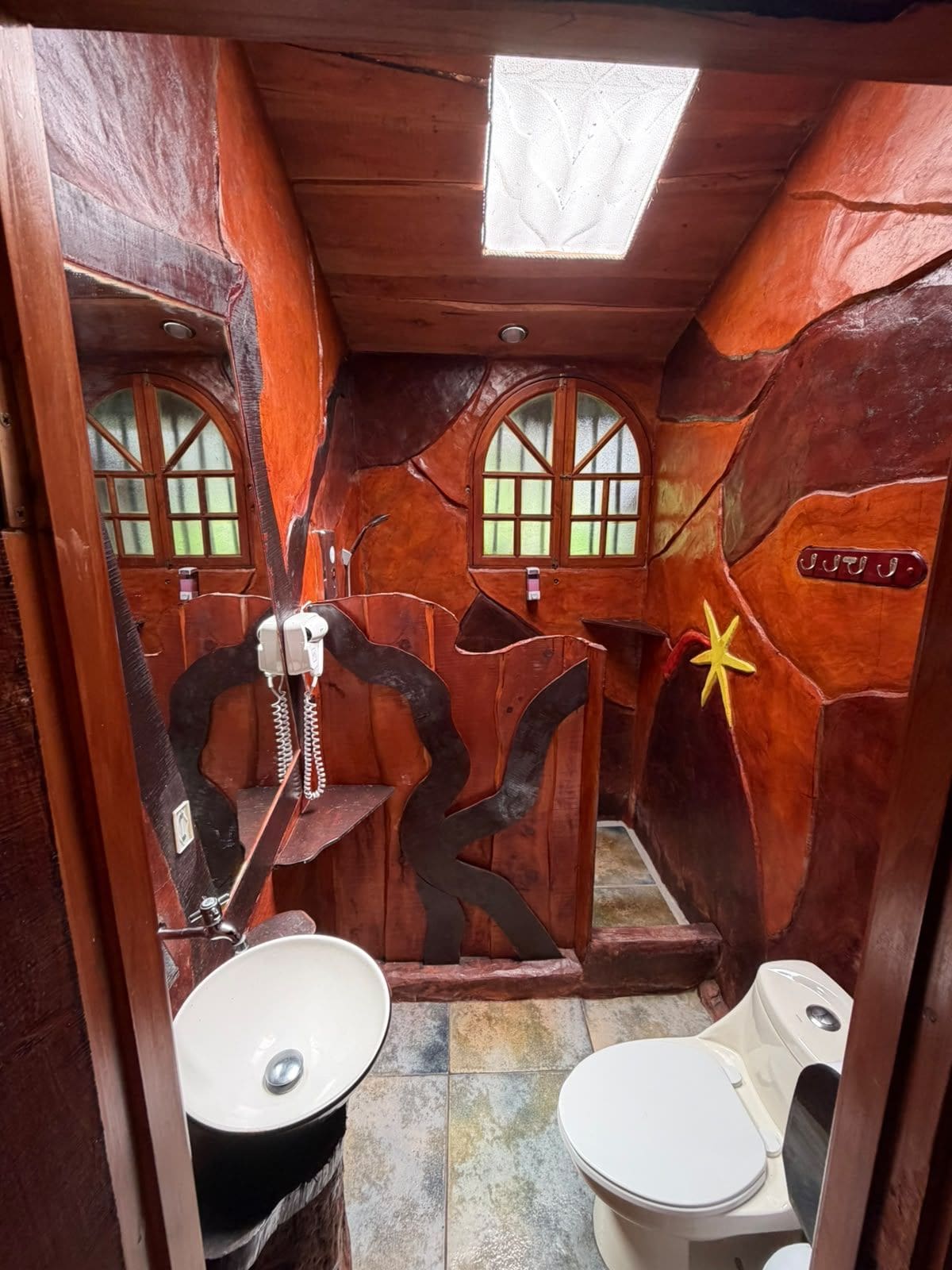 Un baño con diseño rústico, que cuenta con techo de madera, tragaluz, lavamanos, inodoro y una pared decorativa con una estrella.