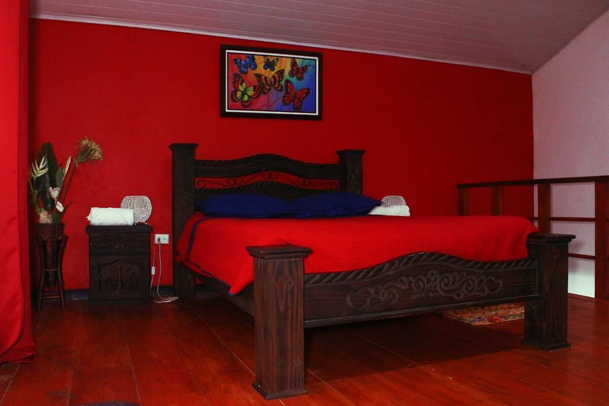 A bedroom with a red wall, a wooden bed with a red bedspread, a blue pillow, and a decorative painting above the bed.