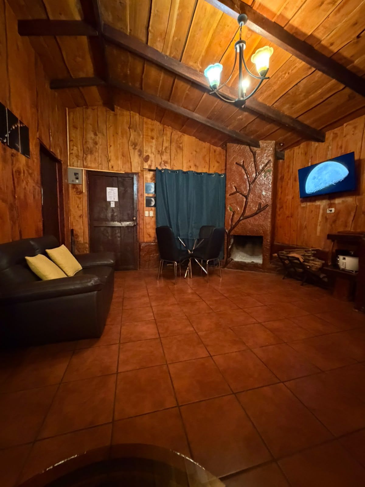A wooden interior with a fireplace, a television mounted on the wall, a black leather sofa, and a tiled floor.