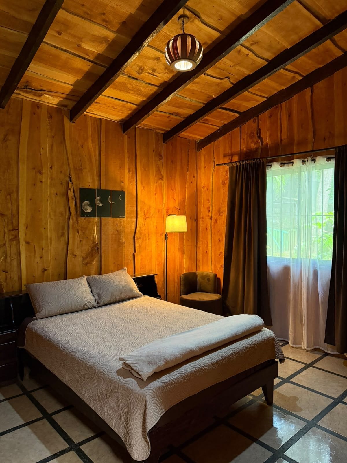 Una stanza in legno con un letto, una sedia, una lampada e una finestra con tende.