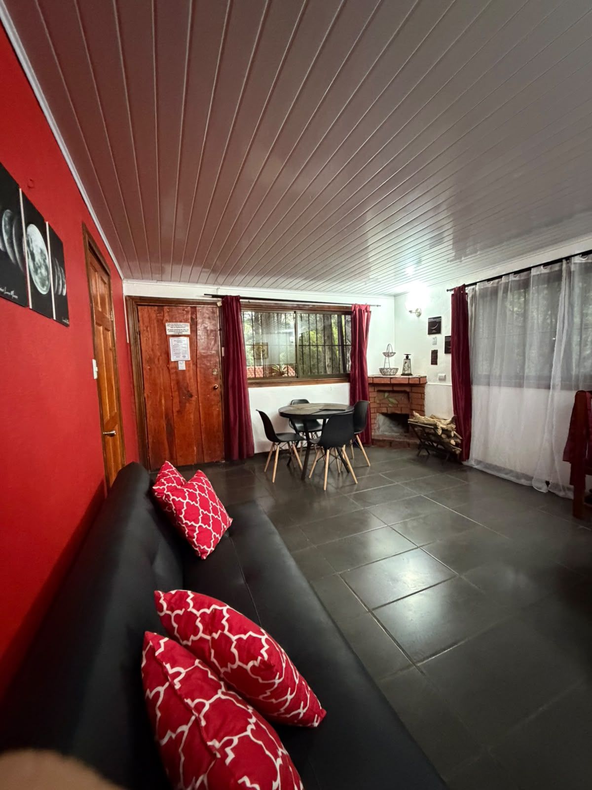 Una sala de estar con un sofá negro, cojines rojos estampados, una mesa de comedor con sillas y vistas al exterior a través de una ventana con cortinas.