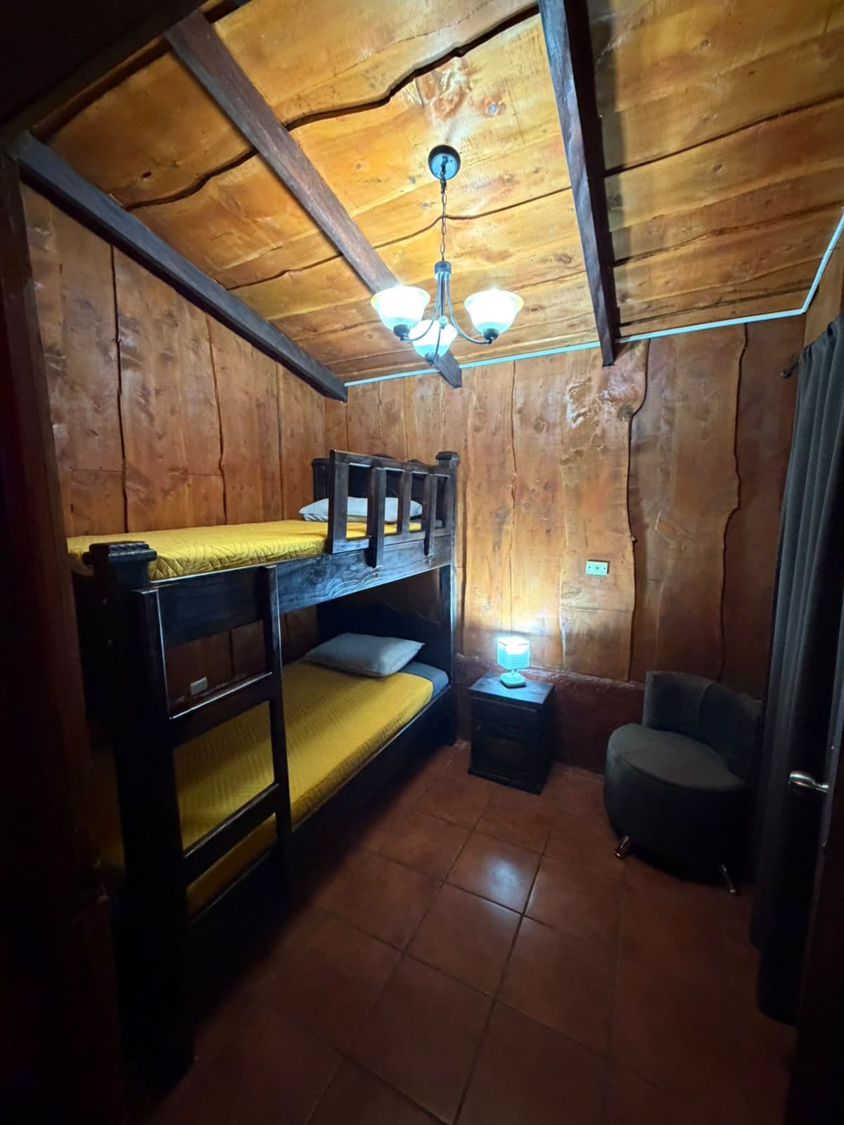 Een kleine, eenvoudige kamer met houten wanden en een schuine kap. Er staan twee bedden: een stapelbed met een gele matras en een eenpersoonsbed met een witte matras. Ook is er een klein tafeltje met een lamp en een stoel.