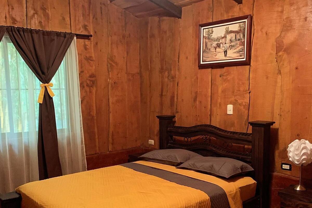 A wooden room with a bed, a nightstand, a lamp, and a framed picture on the wall.