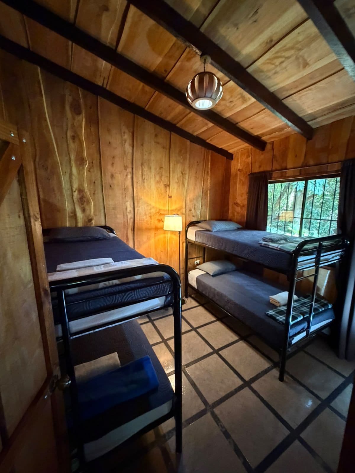 Quarto de cabana de madeira com beliches, uma cama de solteiro, abajur e janela com vista para árvores.