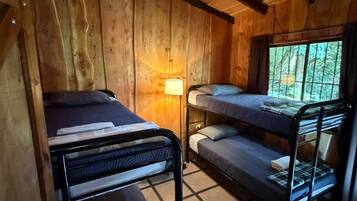 Quarto de cabana de madeira com beliches, uma cama de solteiro, abajur e janela com vista para árvores.
