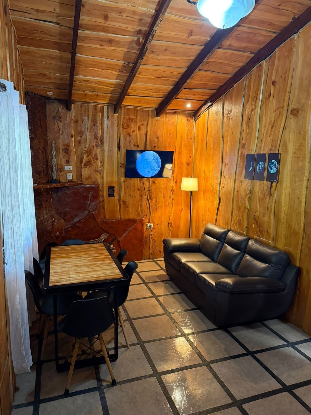 A wooden interior with a dining table, chairs, a sofa, and a wall-mounted TV.