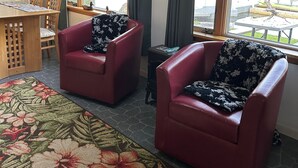 Une pièce comprenant deux fauteuils en cuir rouge, une table à manger en bois avec des chaises, une moquette à motifs floraux et de grandes fenêtres avec des rideaux.