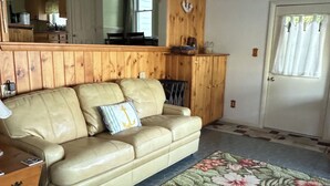 Une pièce avec un canapé en cuir beige, un ventilateur de plafond en bois, un coin cuisine à l’arrière-plan et un tapis à motifs floraux.