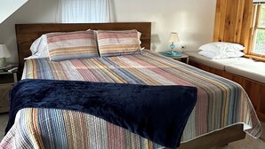 Une chambre avec un lit en bois, une couvre-lit à rayures, un plaid bleu foncé, une table de chevet avec une lampe et une fenêtre avec des volets en bois.
