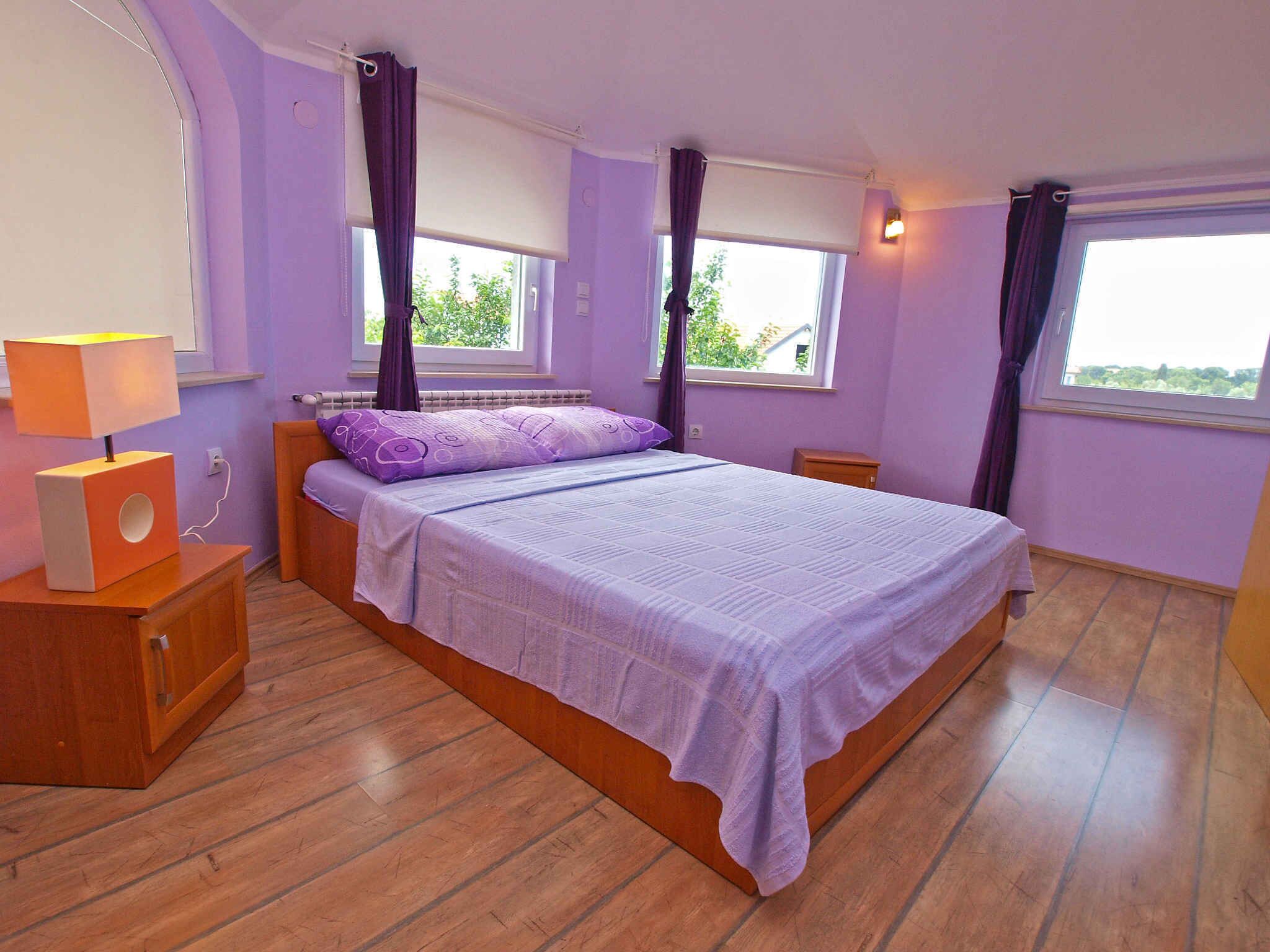 Une chambre à coucher avec un lit, une table de chevet avec une lampe, deux fenêtres avec des rideaux et une vue sur la verdure à l’extérieur.