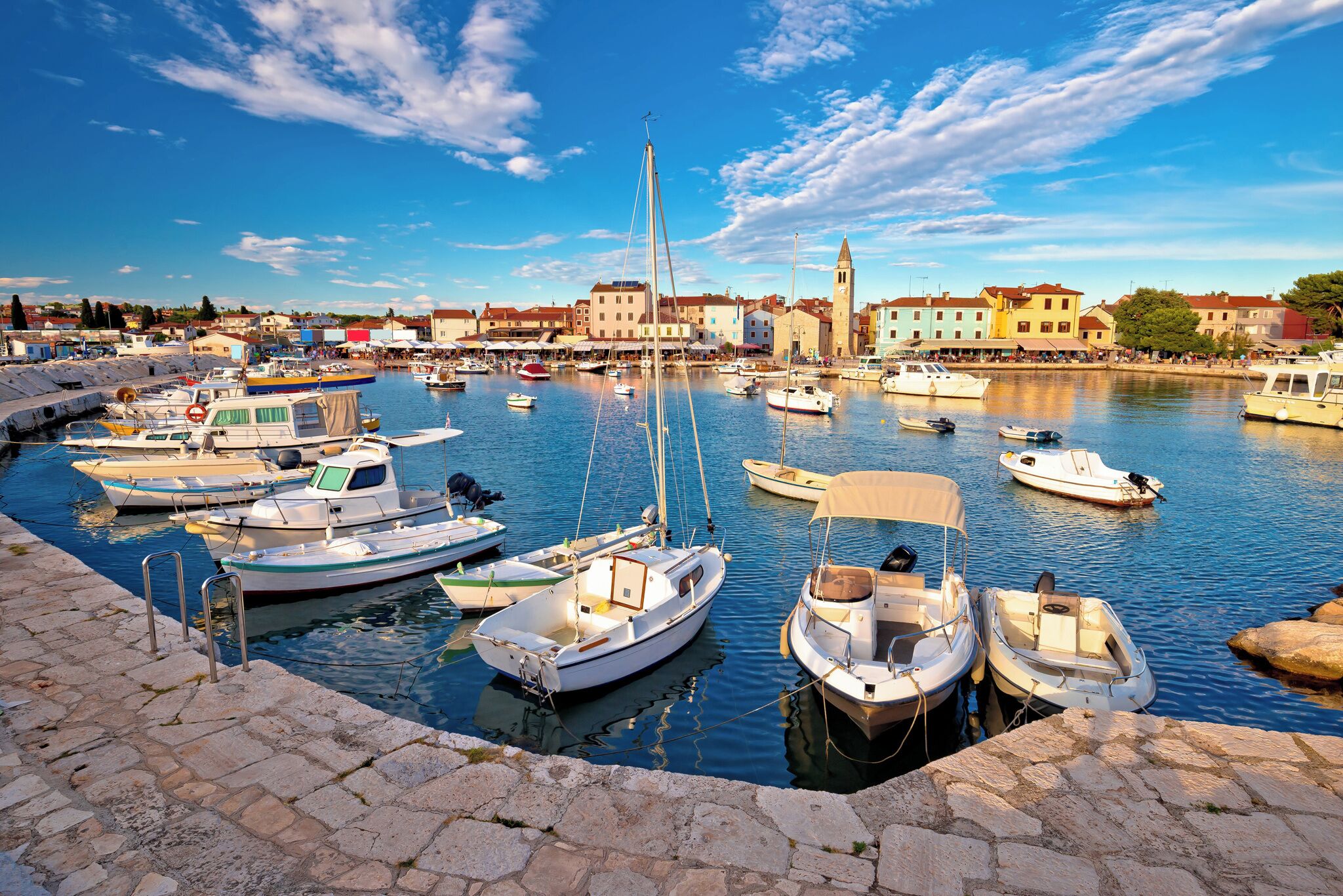 A marina with various boats docked, a stone quay, and buildings in the background.