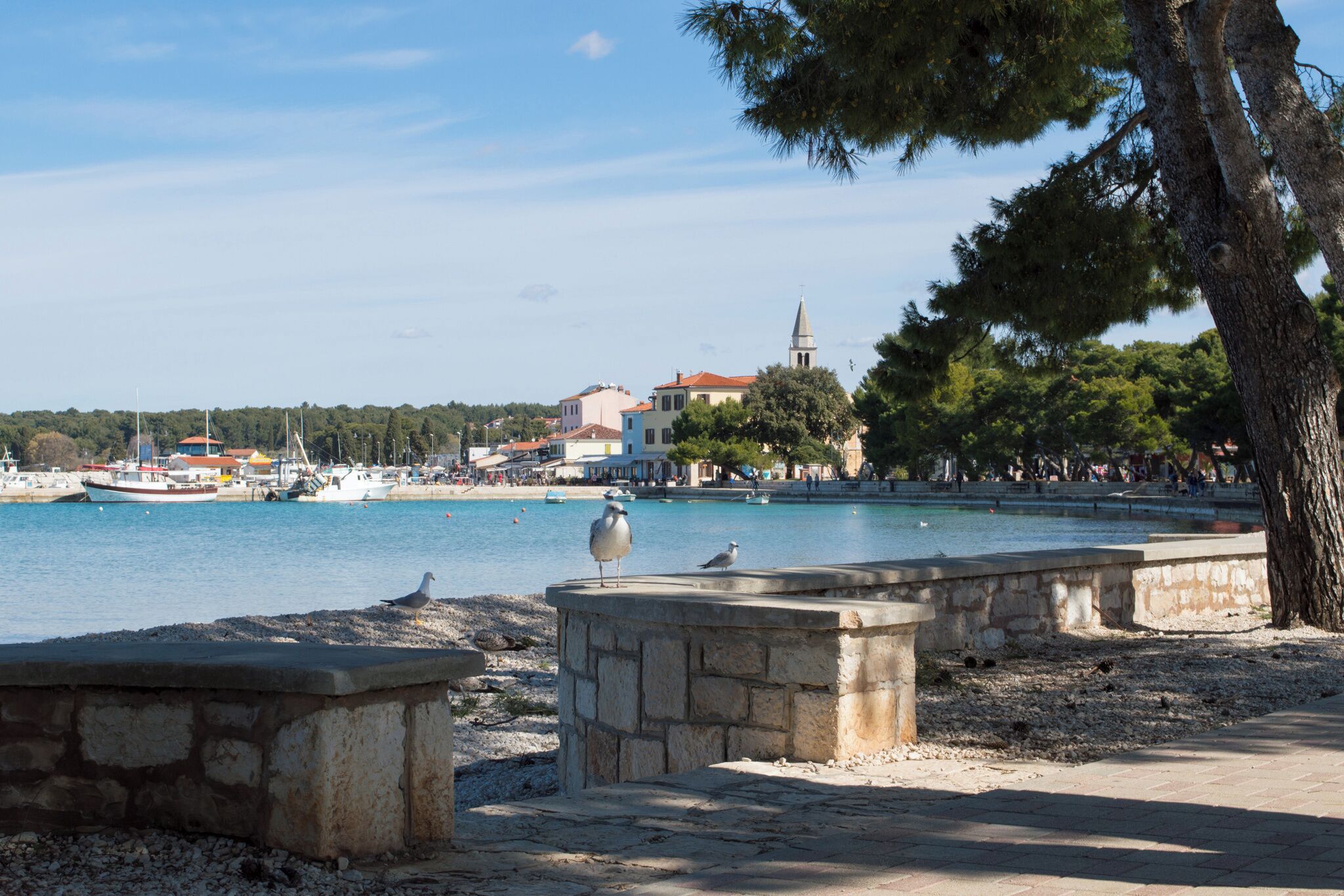 A coastal town with stone benches, a clear blue sea, boats, and a church tower.