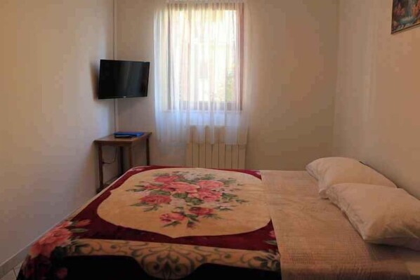 A bedroom with a bed, a television on a stand, a window with curtains, and a radiator.