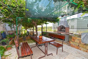 Une terrasse extérieure avec une table et des bancs en bois, entourée de plantes en pot et d’un four en briques.