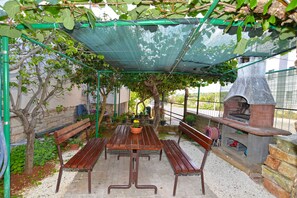 Une terrasse extérieure avec une table et des bancs en bois, un four en briques et une tonnelle recouverte d’un filet vert.