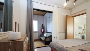 Une chambre à coucher avec un lit, une table de chevet et une commode. Il y a une fenêtre avec des rideaux, un radiateur et une porte qui donne sur une autre pièce.