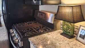 Una cocina con un refrigerador negro, un microondas de acero inoxidable y una estufa. Un gallo está posado sobre el refrigerador, una lámpara decorativa verde sobre la encimera y un cuadro en la pared.