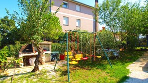 A backyard with a swing set, a barbecue grill, and a table with chairs. - Holiday apartment with garden for shared use and children's playground (Ližnjan)