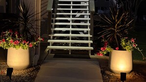 Una escalera con barandilla metálica que lleva a la entrada de un edificio, rodeada de plantas en macetas e iluminación exterior.
