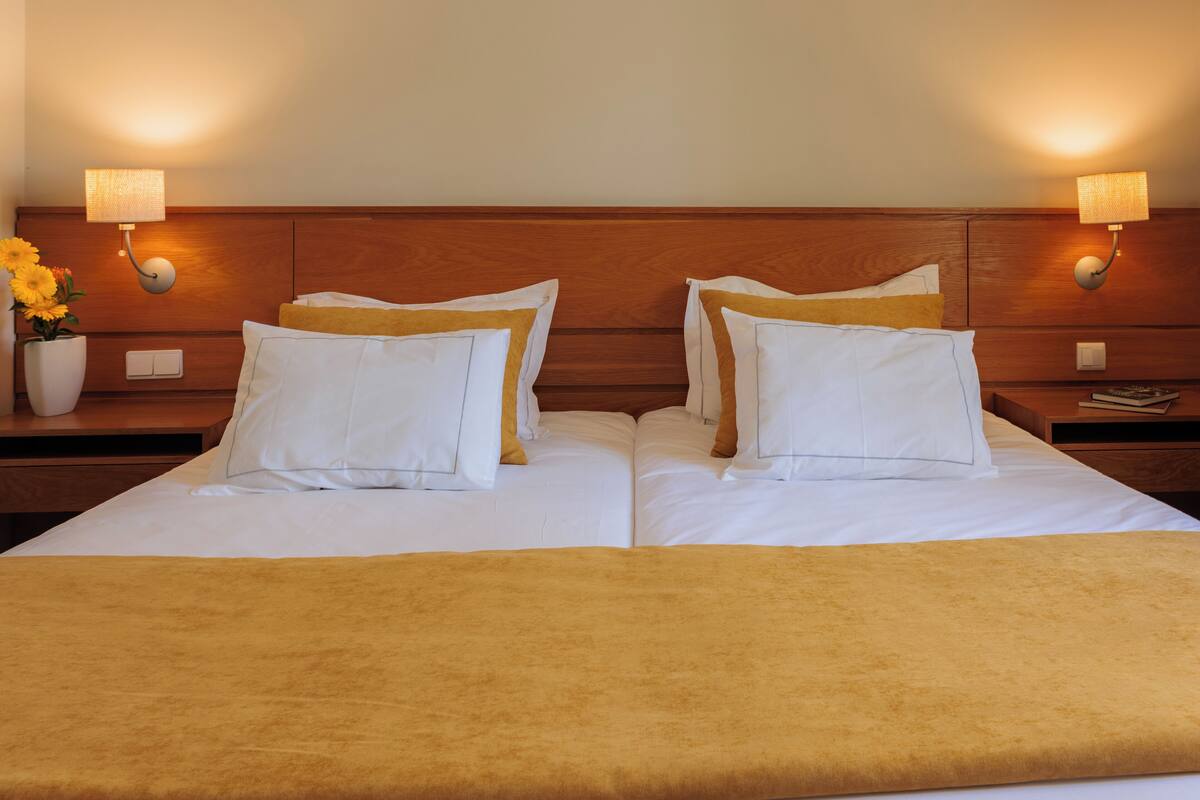 A double bed with white linens and a yellow throw blanket. There are two nightstands with lamps and a vase of flowers.