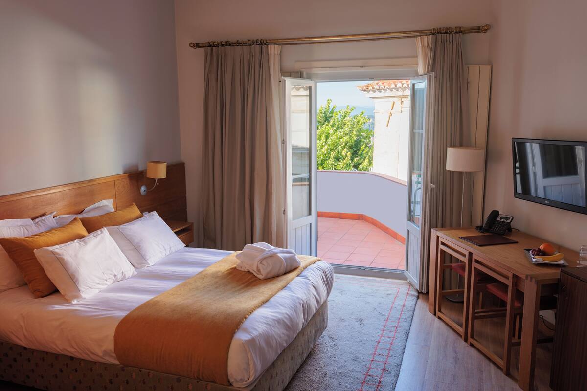 A hotel room with a bed, a desk, a television, and a balcony with a view of greenery.