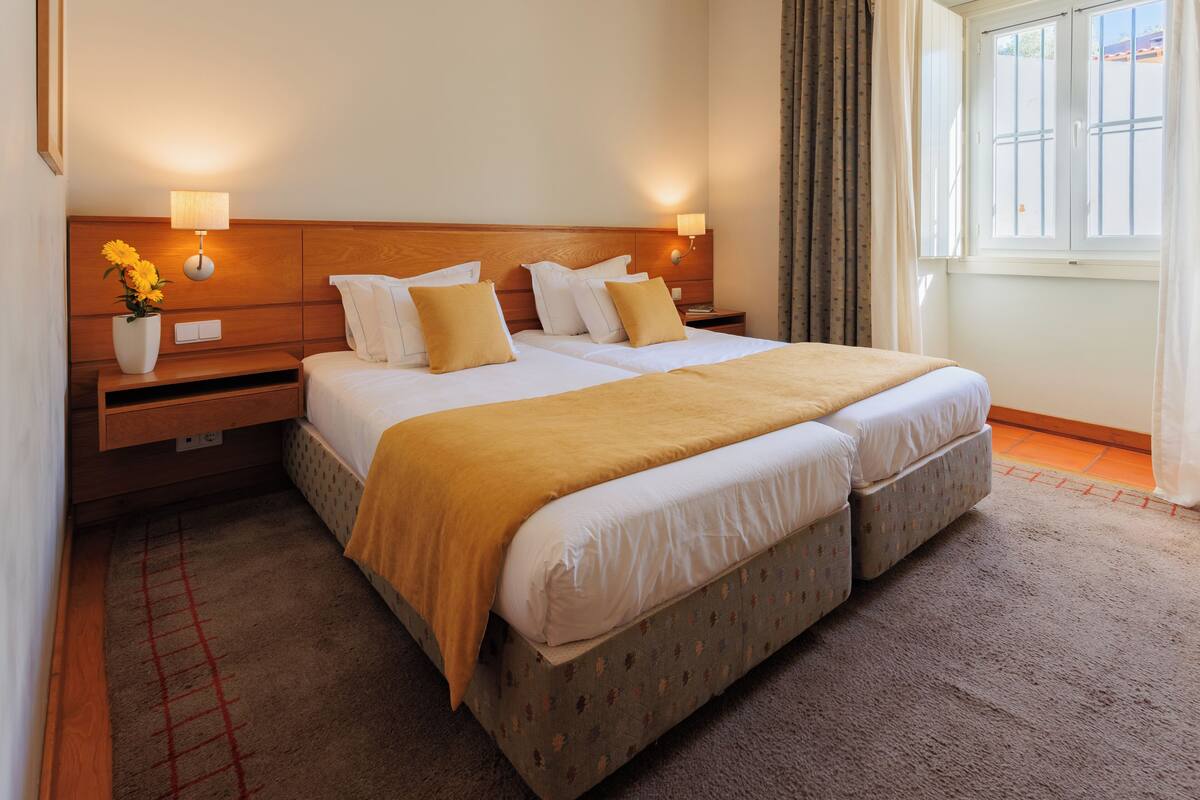 A double bed with a wooden headboard, two bedside tables with lamps, a vase of flowers, and a window with curtains.