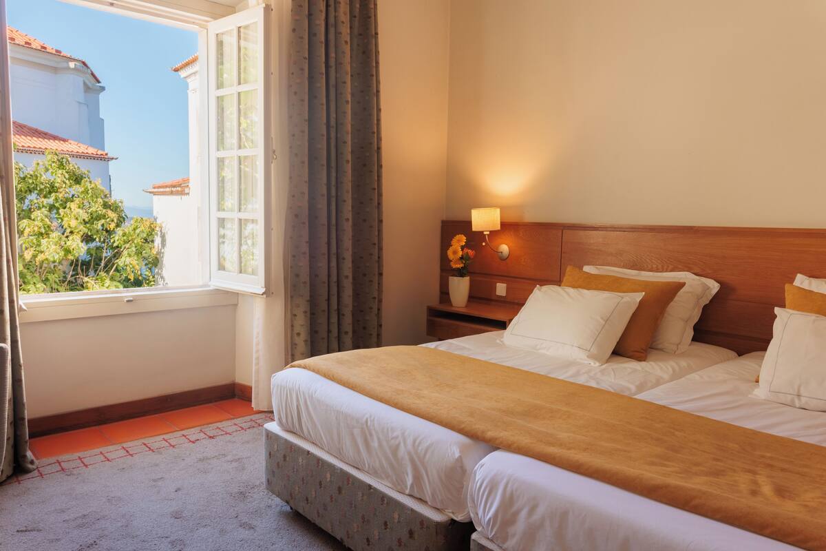 A hotel room with two beds, a window with curtains, a nightstand with a lamp, and a view of buildings outside.