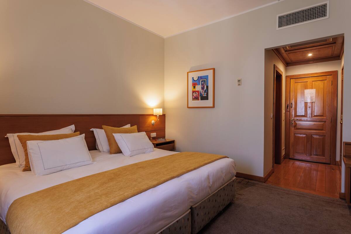 A hotel room with a large bed, a nightstand, a wall-mounted lamp, and a door leading to another room.