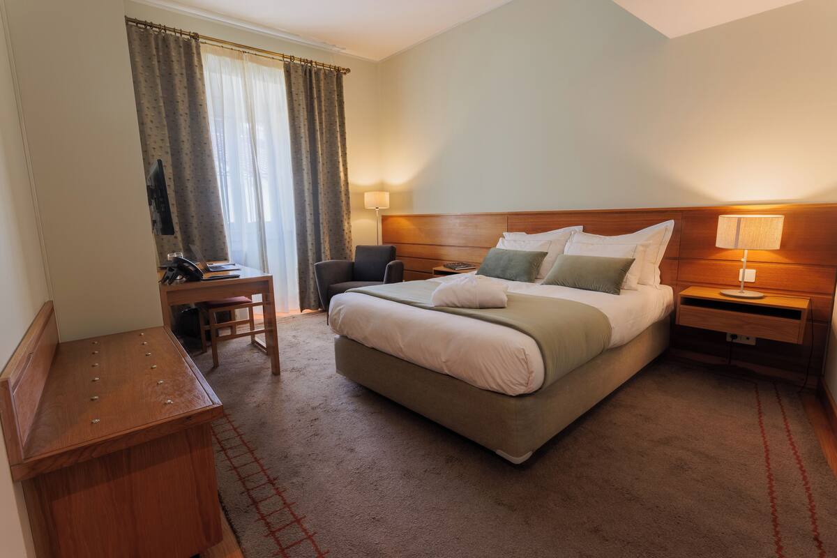 A hotel room with a large bed, a desk with a chair, a TV, and a lamp.
