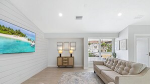 A spacious living room with a large flat-screen TV, a beige sofa, a wooden cabinet, and a view of a beach through a sliding glass door.
