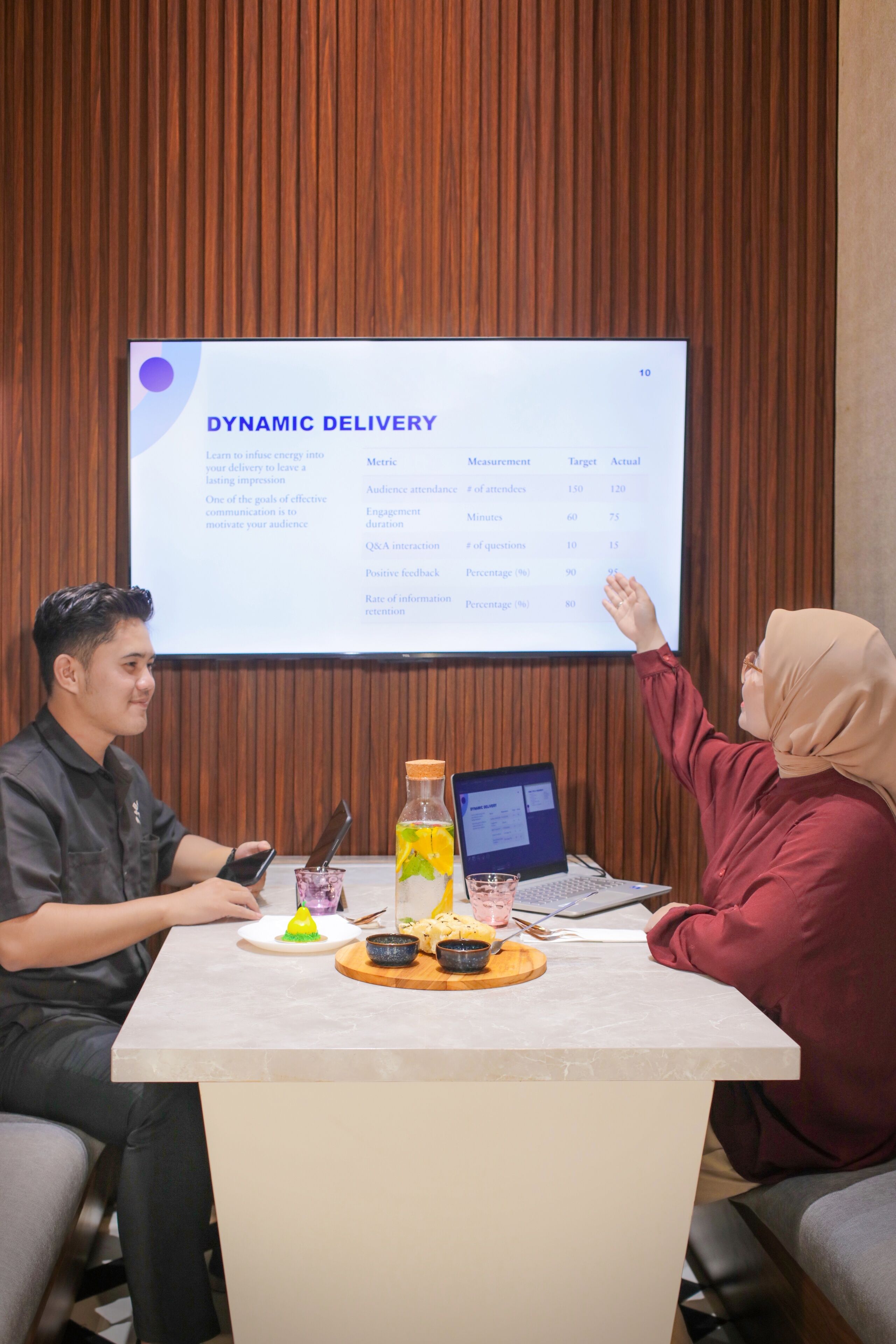 Two people at a table with a presentation screen behind them, a laptop, and a beverage on the table.