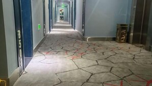 Um corredor com piso rachado, portas azuis e um elevador à direita.