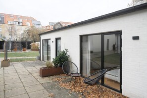 A modern building with large glass doors and windows, a patio with chairs, and a garden area with plants.