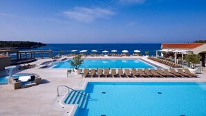 A resort pool area with two large pools, sun loungers, umbrellas, and a view of the sea.