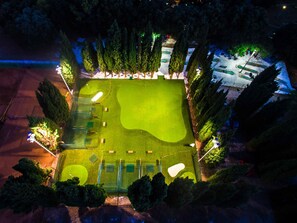 An aerial view of a golf course at night with well-lit areas, surrounded by tall trees and a parking lot.