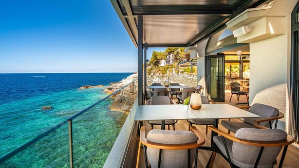 A balcony with a table and chairs overlooking a coastal area with clear blue water and rocky shore.
