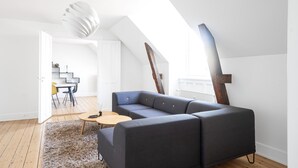 Egy modern, loft stílusú lakás, melyben egy nagy, szürke, moduláris kanapé, egy kerek, fából készült dohányasztal és egy puha szőnyeg található. Egy lépcső vezet a felső szintre, a mennyezetről pedig egy különleges, fehér, függesztett lámpa lóg alá.