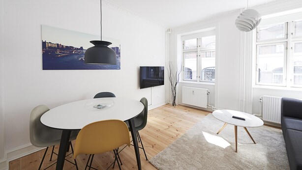 A modern living room with a round dining table, a sofa, and a large window with a view of buildings. - Nice 1-bed Near Copenhagen Lakes (Copenhagen)