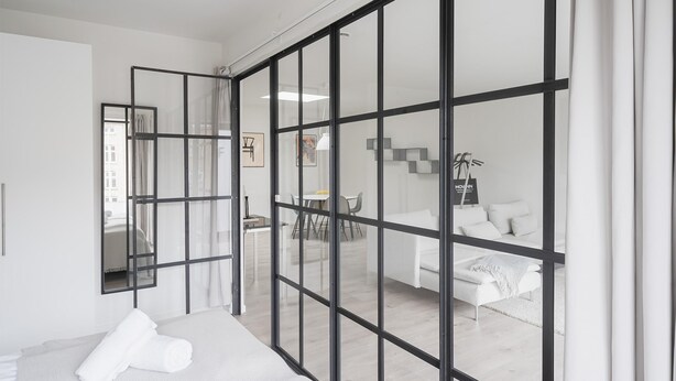 Une chambre d’hôtel moderne dotée d’une grande porte-fenêtre coulissante, d’un lit avec du linge de maison blanc, d’une petite table avec une chaise et d’un canapé dans l’espace salon attenant.