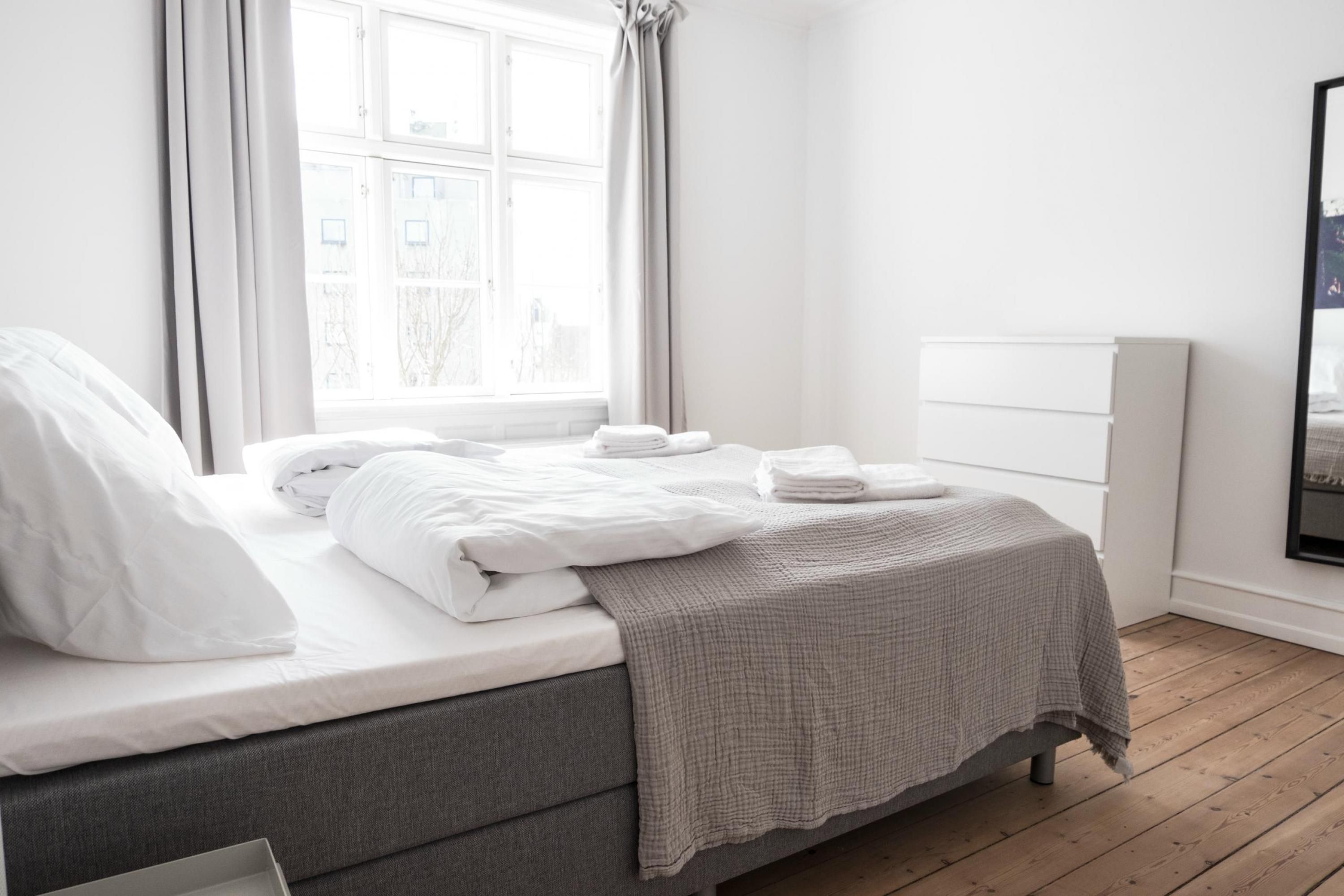 A neatly made bed with white linens and a grey blanket, a window with curtains, and a white wall with a radiator.